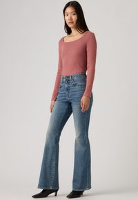 Μπλούζα με μακριά μανίκια και στρογγυλή λαιμόκοψη σε χρώμα σκουριασμένο ροζ, συνδυασμένη με ψηλόμεσα, καμπάνα μπλε τζιν, με ξεθωριασμένο φινίρισμα, και σκοτεινές μπότες με τακούνι.