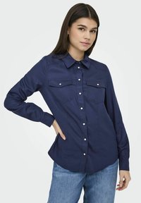 Camicia blu navy con colletto, due tasche sul petto e vestibilità classica. Presenta bottoni bianchi e maniche lunghe con polsini abbottonati.