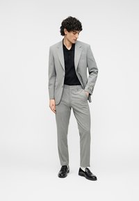 Φωτεινό γκρι κοστούμι με σακάκι και παντελόνια ραμμένα στα μέτρα. Μαύρο πόλο πουκάμισο από μέσα. Μαύρα παπούτσια συμπληρώνουν την εμφάνιση. Διακριτικό ριγέ μοτίβο.