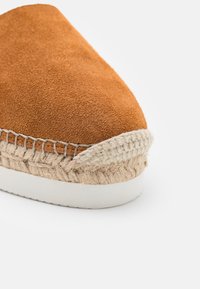 Gros plan sur une espadrille en daim marron avec des détails en jute tissé et une semelle en caoutchouc blanc sur fond blanc.