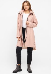 Ljusrosa vattentät jacka med huva, med en framsida med dragkedja, tryckknappar, sidofickor och justerbara ärmar, kombinerad med svarta byxor och stövlar.