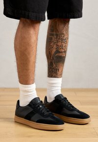 Black suede and synthetic sneakers with a gum sole, featuring textured panels and lacing. White ribbed socks worn above the ankle.