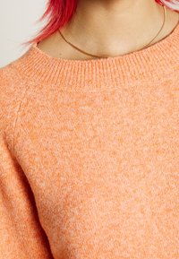 Suéter de punto suave y texturizado, con un cuello redondo acanalado. El color es naranja claro con variaciones sutiles en todo su tejido.