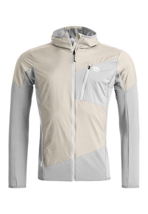 Person, die den Ärmelbund eines hellgrauen, langärmligen Sportshirts justiert, trägt eine beige Weste und blaue Hose.