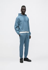 Blauwe hoodie met verstelbare trekkoorden, gecombineerd met bijpassende blauwe joggingbroek. Simpel ontwerp, zachte textuur en een comfortabele pasvorm. Zwarte schoenen.
