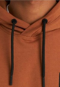 Hnědá mikina s kapucí a černými šňůrkami, kterou nosí osoba, s detailním prošíváním kolem kapuce a výstřihu.