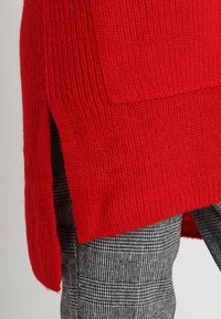 Pull rouge en maille avec un motif texturé et des fentes latérales, associé à un pantalon à carreaux noirs et blancs présentant un tissu structuré.