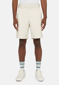 Pantaloni corti beige su misura con frontale piatto, passanti per cintura e tasche laterali. Indossati con calze a righe verdi e bianche e sneaker bianche.
