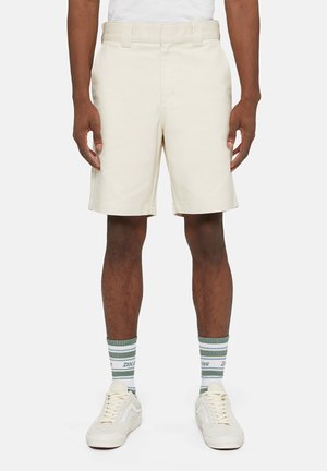 Pantaloni corti beige su misura con frontale piatto, passanti per cintura e tasche laterali. Indossati con calze a righe verdi e bianche e sneaker bianche.