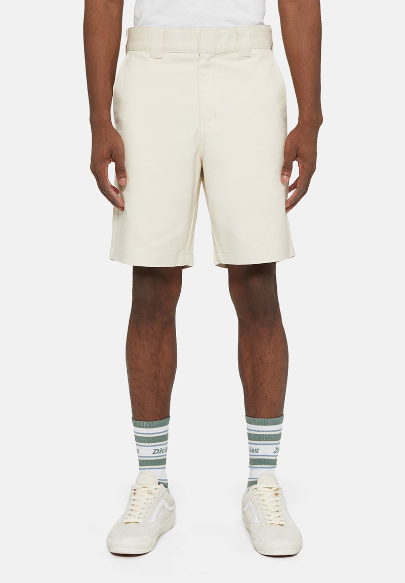 Pantaloni corti beige su misura con frontale piatto, passanti per cintura e tasche laterali. Indossati con calze a righe verdi e bianche e sneaker bianche.