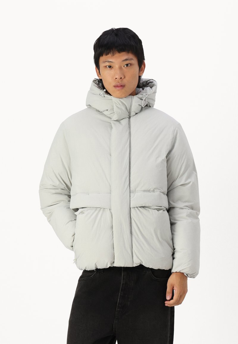 Chaqueta acolchada gris claro con un alto cuello, bolsillos frontales y un dobladillo ajustado. Presenta una textura suave y mate, además de una capucha ajustable.