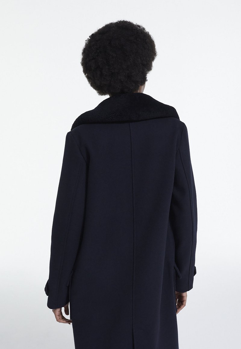 Long manteau bleu marine avec un col en velours noir. Présente une coupe droite, des fentes latérales et des poignets boutonnés. Texture lisse et design minimaliste.