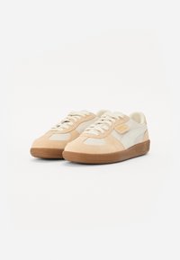 Beige sporta apavi ar zamšas un tekstila augšdaļu, baltajiem akcentiem, gumijas zoli, apaļu purngalu un klasisku šņorēšanas dizainu. Zelta logotipa detaļa sānos.