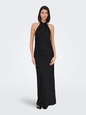 Jeune femme aux cheveux raides et foncés portant une robe de soirée noire sans manches à col halter, debout devant un fond clair uni.