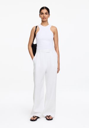 Vrouw met een witte mouwloze top en witte broek met wijde pijpen, met een zwarte schoudertas, staand tegen een effen witte achtergrond.