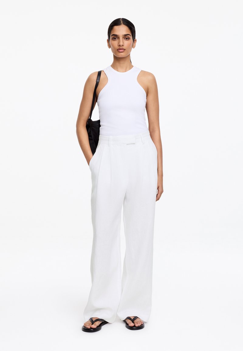 Femme portant un haut blanc sans manches et un pantalon blanc à jambes larges, tenant un sac à bandoulière noir, debout devant un fond blanc uni.
