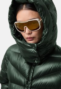 Grøn pufjakke med en skinnende finish, der har en høj krave og en stor hætte. Accentueret af hvide oversized solbriller med gule linser.
