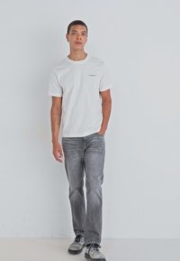 Hvit bomull T-skjorte med korte ermer og en liten logo. Grå denimjeans med rette ben. Mørke sko med snøring.