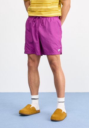 Person iført lyse lilla Nike-shorts, hvide sokker med sorte striber, sennepsgule slip-on sko og en gulstribet skjorte, stående på blå gulv.