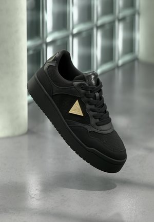 Black sneakers with textured leather, a gold triangular logo, and round laces. The sole is thick and rubberized, with a flat profile.