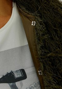 Brown outer garment with textured, fringed trim. Layered over a white shirt featuring a black and white graphic print. Silver hardware accents.