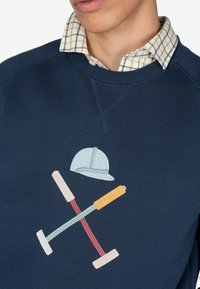 Marineblå sweatshirt med et farverigt broderet design af krydsede mallets og en hjelm. Ribbet halsudskæring og skjortekrave i tern er synlig.