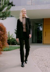 Lächelnde Frau mit langen blonden Haaren, die auf einem Weg neben Gartenpflanzen geht, trägt ein schwarzes Blazer, einen braunen Rollkragenpullover, schwarze Hose und Stiefel.