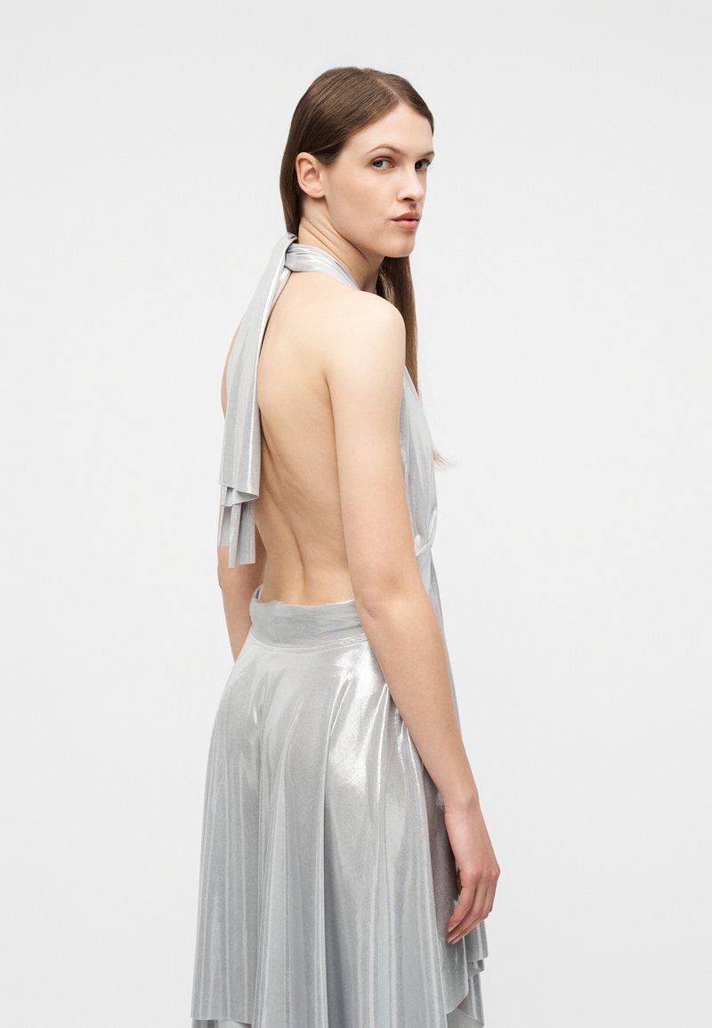 Femme aux longs cheveux bruns portant une robe argentée dos nu à col halter, regardant par-dessus son épaule sur un fond uni clair.