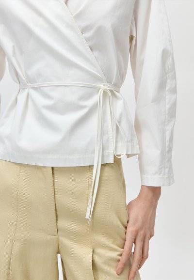 Biała, długa bluzka z kopertowym zapięciem, wiązana z boku, zestawiona z jasnobeżowymi teksturowanymi spodniami, widoczna od połowy tułowia do rąk.