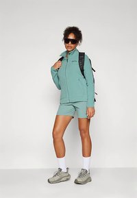 Donna vestita con una giacca a zip color teal e pantaloni corti, calze bianche, scarpe da trekking grigie, zaino nero e grandi occhiali da sole neri che si trova su uno sfondo neutro.