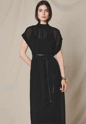 Kobieta stojąca na jednolitym tle, ubrana w prześwitującą czarną suknię z wysokim kołnierzem, paskiem w talii i złotymi bransoletkami na nadgarstku.