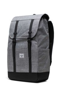 Mochila de tela gris con correas negras, cierre con solapa, bolsillo frontal y un diseño texturizado. Presenta una etiqueta de marca en el frente.