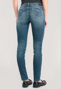 Jean skinny en denim bleu moyen, avec une taille haute, des poches arrière avec des motifs angulaires et un léger effet de décoloration, associés à des chaussures noires.