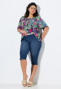 Sininen, kukkakuosisessa lyhythihaisessa paitapuserossa, yhdistettynä tummiin farkkushortseihin ja beigeihin liukuviin sandaaleihin, joissa on teksturoitu pinta.