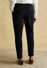 Tumši zili korduroveļa bikses ar piegulošu dizainu un vidēja augstuma jostasvietu, divām kabatām aizmugurē un smalku faktūru, kopā ar brūniem apaviem.