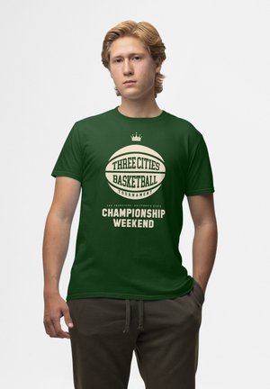 Camiseta de algodón verde con un gráfico central de baloncesto y texto. El diseño incluye "Torneo de Baloncesto de Tres Ciudades" y "Fin de Semana de Campeonato."