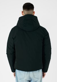 Giacca nera con cappuccio caratterizzata da una texture imbottita, con silhouette aderente e polsini elasticizzati. Vista posteriore, indossata con jeans azzurri chiari.