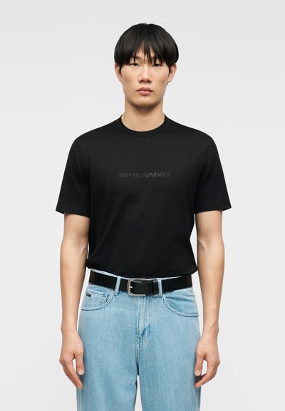 Νέος άνδρας που φοράει μαύρο μπλουζάκι Emporio Armani, φορεμένο μέσα σε ανοιχτόχρωμο μπλε τζιν με μαύρη ζώνη, στέκεται μπροστά σε απλό φόντο.