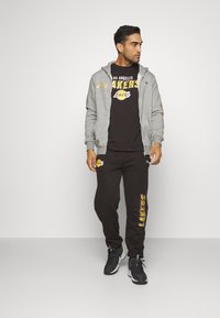 Sudadera gris con cremallera y logo de Los Angeles Lakers, camiseta negra debajo, pantalones de chándal negros con estampado de Lakers y zapatillas deportivas negras.