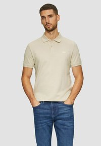 Beige Poloshirt mit Kragen, kurzen Ärmeln und dezentem Logo. Trägt dazu eine blaue Jeans, das Modell steht mit den Händen in den Taschen.