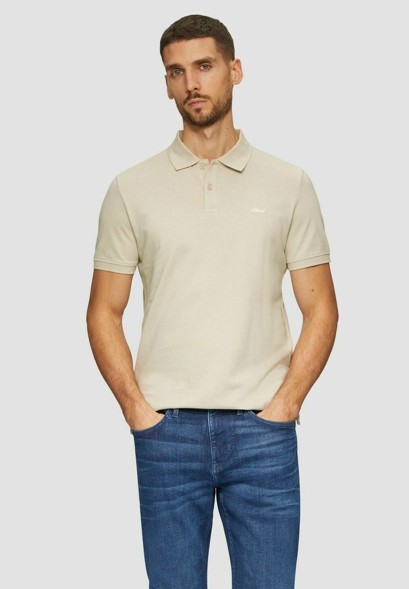 Beige Poloshirt mit Kragen, kurzen Ärmeln und dezentem Logo. Trägt dazu eine blaue Jeans, das Modell steht mit den Händen in den Taschen.
