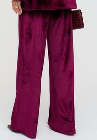 Plüschige, weit geschnittene Hosen aus samtigem, tiefburgunderrotem Samt. Der Stoff hat eine glatte Textur, reflektiert das Licht und weist ein locker sitzendes, fließendes Design auf.