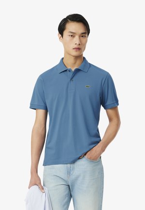 Joven con una camisa polo azul con un pequeño logo de cocodrilo sosteniendo una prenda blanca, jeans de lavado claro, de pie contra un fondo blanco.