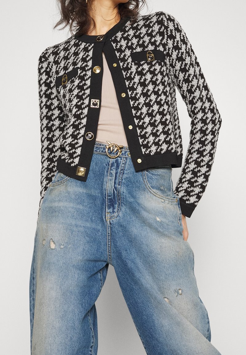 Femme portant un cardigan pied-de-poule noir et blanc avec des boutons dorés, un top beige, une ceinture à boucle dorée et un jean bleu délavé et ample avec des déchirures.