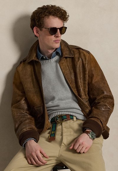Man met krullend haar, zonnebril, bruine leren jas, grijze trui, blauw overhemd, gestreepte riem, beige broek en polshorloge, zittend tegen een eenvoudige muur.