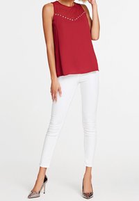Top rosso senza maniche con scollatura trasparente e borchie decorative; abbinato a jeans skinny bianchi e scarpe con tacco metalliche.