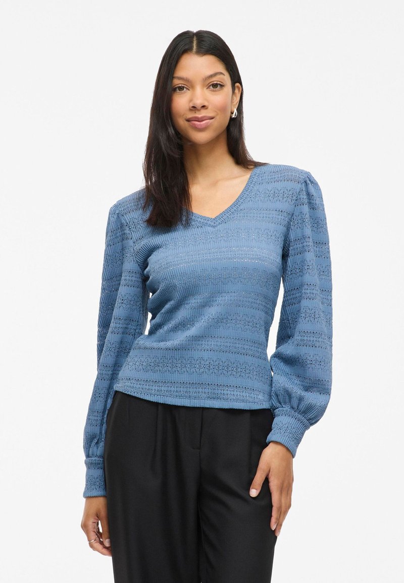 Blauwe gebreide trui met een V-hals, een gestructureerd patroon en opgeblazen lange mouwen, gecombineerd met zwarte hoog-getailleerde broek.