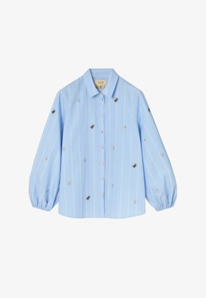 Világoskék, gombos blúz fehér, függőleges csíkokkal, hosszú, puffadt ujjakkal, valamint apró hímzett virág- és méhmintákkal az ujjakon és a testen szétszórva.