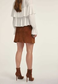 Biała bluzka z warstwowymi falbanami, brązowa zamszowa mini spódniczka, brązowe botki na obcasie w stylu stiletto. Gładka faktura, dopasowany krój.