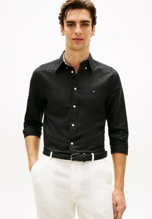 Νέος άνδρας που φοράει μαύρο πουκάμισο με κουμπιά και μανίκια γυρισμένα, λευκό παντελόνι και μαύρη πλεκτή ζώνη, στέκεται με τα χέρια στις τσέπες.